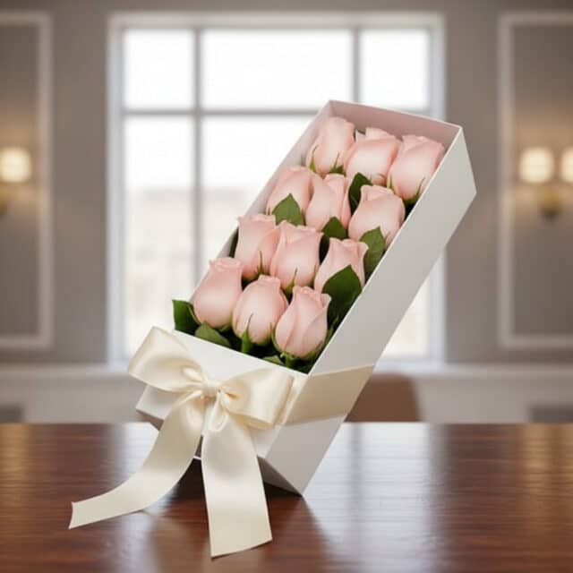 Caja blanca inclinada con 12 rosas rosadas frescas y un moño de satén, sobre una mesa de madera oscura con un fondo elegante