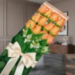Caja de 12 rosas salmón frescas con follaje verde y alstroemerias blancas, presentadas en un empaque transparente con lazo be