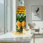 Caja de 18 rosas amarillas frescas con alstroemerias blancas, lazo dorado, tarjeta de regalo y cinta de satén sobre mesa de m