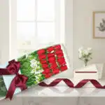 Caja de 18 rosas rojas frescas con alstroemerias blancas, lazo de satén burdeos y tarjeta de regalo en sobre, sobre mármol.