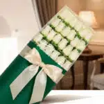 Caja de 24 rosas blancas frescas y elegantes, envueltas en papel verde oscuro con un moño de satén color crema, sobre un fond