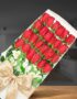 Caja de 24 rosas rojas frescas con gotas de rocío, lazo dorado y flores blancas de acompañamiento, sobre un fondo elegante de