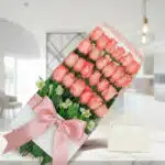 Caja de 24 rosas rosadas frescas con lazo de satén y tarjeta de regalo, sobre mármol en un ambiente elegante.