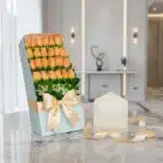 Caja de 24 rosas salmón frescas en empaque azul con lazo satinado, tarjeta de regalo y cinta sobre mármol.