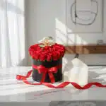 Caja redonda negra con 25 rosas rojas frescas y una orquídea blanca, adornada con una cinta de satén rojo, sobre una mesa de
