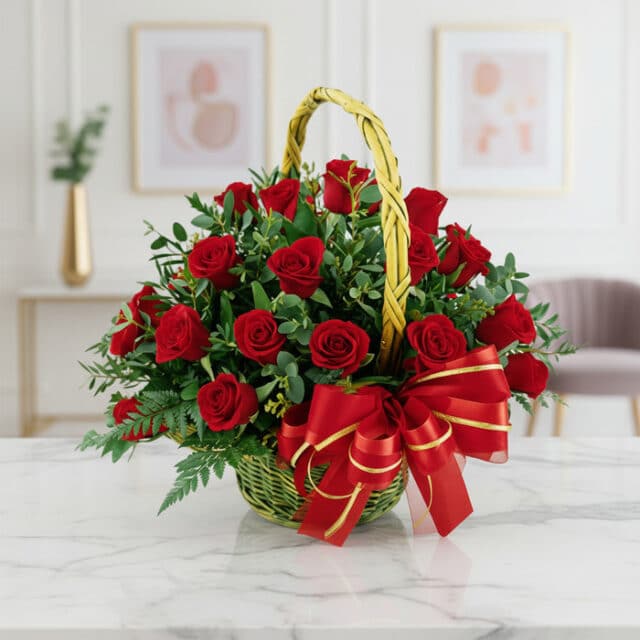 Canasta floral de mimbre verde con 24 rosas rojas vibrantes, follaje verde y un lazo rojo brillante, sobre una mesa de mármol