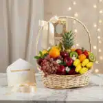 Canasta de mimbre con frutas frescas variadas como uvas, manzanas, naranjas, kiwis, fresas, plátanos y piña, decorada con un