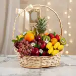 Canasta de mimbre con frutas frescas variadas como piña, uvas, manzanas, kiwis, naranjas, peras, fresas y plátanos, decorada