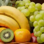 Primer plano de una canasta de frutas Corvus con plátanos, uvas verdes, kiwi, naranja y manzana, destacando su frescura y col