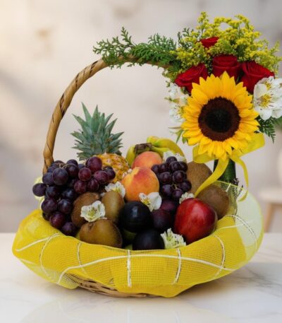 Canasta de Frutas Draco con uvas, manzanas, duraznos, kiwis, piña y arreglo floral de rosas, girasol y flores blancas sobre m