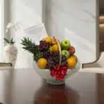 Canasta de frutas gourmet con piña, uvas, manzanas y naranjas, sobre una mesa de madera pulida en un entorno elegante.