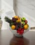 Canasta de frutas gourmet con piña, uvas, manzanas y naranjas, sobre una mesa de madera pulida en un entorno elegante.