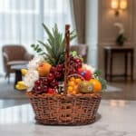 Canasta de mimbre con piña, uvas rojas, manzanas, duraznos, kiwis, naranjas y plátanos, con flores blancas y eucalipto, sobre