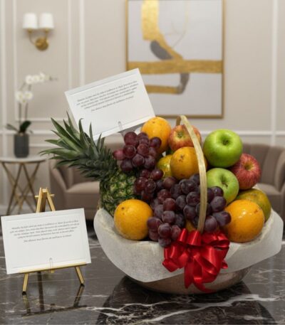 Canasta de frutas premium con piña, uvas, manzanas rojas y verdes, naranjas, y un lazo rojo, sobre una mesa de mármol en un e