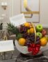 Canasta de frutas premium con piña, uvas, manzanas rojas y verdes, naranjas, y un lazo rojo, sobre una mesa de mármol en un e