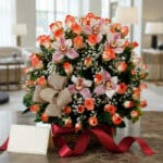 Canasta de rosas y orquídeas en tonos salmón y rosa, con gypsophila y un lazo de yute, sobre una mesa de mármol con una cinta