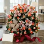 Canasta de rosas y orquídeas en tonos salmón y rosa, con gypsophila y un lazo de yute, sobre una mesa de mármol con una cinta