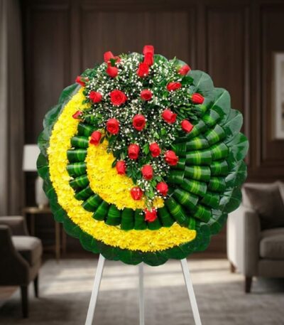 Corona fúnebre Adela con rosas rojas y flores blancas sobre base de crisantemos amarillos y hojas verdes, en sala de velación