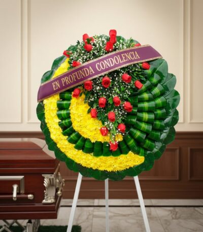Corona fúnebre Adela con rosas rojas y flores amarillas, cinta morada 'En Profunda Condolencia', junto a un ataúd en sala de