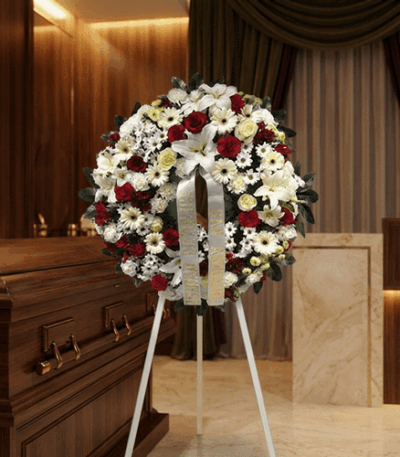 Corona de flores fúnebres Ararat con rosas rojas, lirios blancos y margaritas, junto a un ataúd de madera en un ambiente eleg