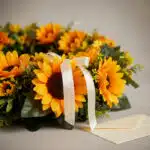 Fotografía macro premium de una corona fúnebre con girasoles vibrantes, follaje verde y una cinta satinada blanca. Los pétalo