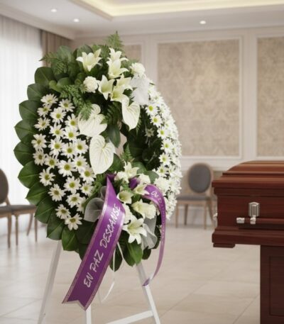 Corona fúnebre Legado de Paz con flores blancas, liliums, margaritas y anturios, cinta morada 'En Paz Descanse' en sala de ve