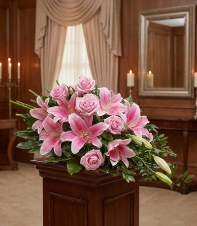 Corona fúnebre Lino con rosas rosadas y lirios, exhibida elegantemente en una sala de velación con iluminación cálida y fondo