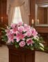Corona fúnebre Lino con rosas rosadas y lirios, exhibida elegantemente en una sala de velación con iluminación cálida y fondo