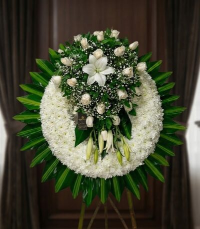 Corona fúnebre grande con rosas blancas, lirios y follaje verde, presentada en una sala de velación premium con luz natural s