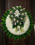 Corona fúnebre grande con rosas blancas, lirios y follaje verde, presentada en una sala de velación premium con luz natural s