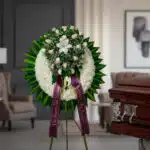 Impresionante corona fúnebre con rosas blancas, lirios y follaje verde, sobre un caballete, con cinta morada de condolencia,
