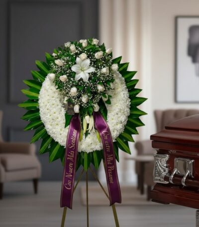 Impresionante corona fúnebre con rosas blancas, lirios y follaje verde, sobre un caballete, con cinta morada de condolencia,
