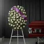Corona fúnebre de rosas blancas y follaje verde, con una cinta morada que dice 'En memoria cariñosa', junto a un ataúd de mad