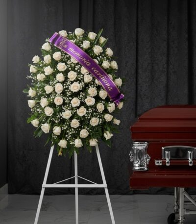 Corona fúnebre de rosas blancas y follaje verde, con una cinta morada que dice 'En memoria cariñosa', junto a un ataúd de mad
