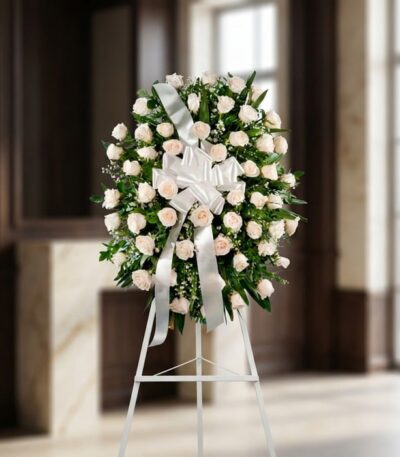 Corona fúnebre Serenidad con rosas blancas y follaje verde, lazo blanco, sobre un trípode en una sala de velación premium con