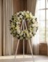 Corona fúnebre "Solemne Homenaje Joshua" con lirios blancos, rosas y crisantemos, sobre un caballete en una sala de velación