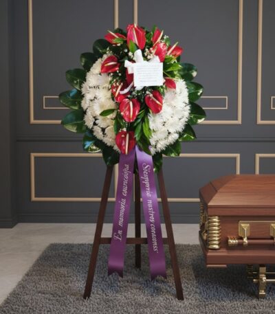Corona fúnebre Trinidad con anthuriums rojos y crisantemos blancos, cinta morada con mensaje de condolencia, junto a un ataúd