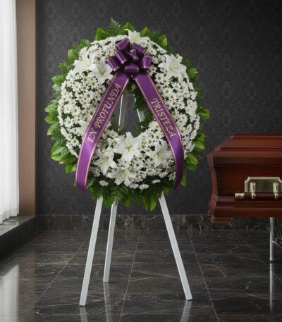 Corona de homenaje "El Vuelo de Uriel" con flores blancas y cinta morada con mensaje de condolencia, junto a un ataúd en sala