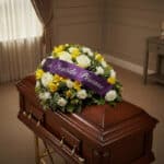 Cubre caja fúnebre Baltasar con rosas blancas, fresias amarillas y follaje verde, adornado con una cinta morada que dice 'Con