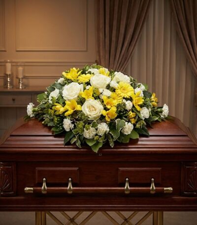 Cubre Caja Fúnebre Baltasar con rosas blancas, freesias amarillas y margaritas, sobre un ataúd de madera oscura en un entorno