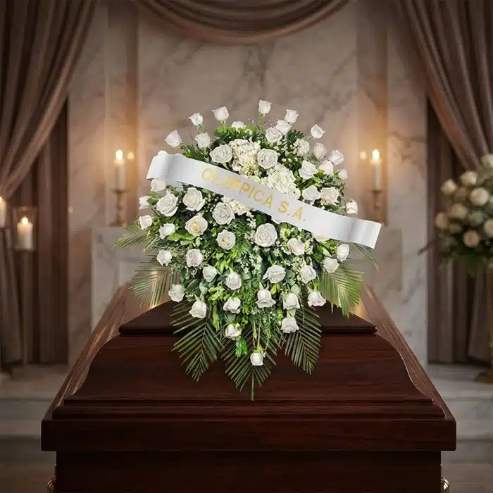 Cubre Caja Fúnebre Bernardita: Un Manto de Paz y Amor Cubre caja fúnebre Bernardita con rosas blancas y follaje verde sobre un ataúd de madera oscura, en un entorno elegante y sol