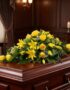 Elegante cubre caja fúnebre con rosas amarillas y lirios, sobre un ataúd de madera oscura en un ambiente solemne y bien ilumi