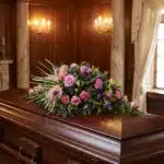 Cubre caja fúnebre 'Legado Sereno de Ivon' con rosas rosadas, claveles y cardos azules sobre un ataúd de madera oscura en un