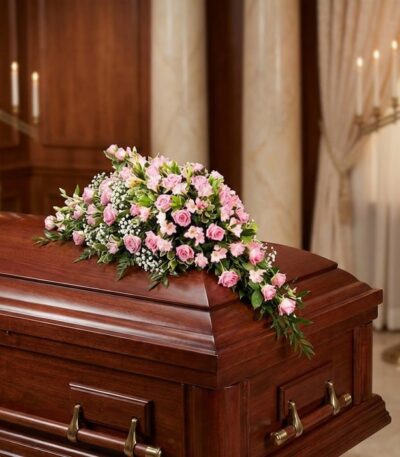 Cubre caja fúnebre elegante con rosas rosadas y follaje verde, delicadamente dispuesto sobre un ataúd de madera oscura en un