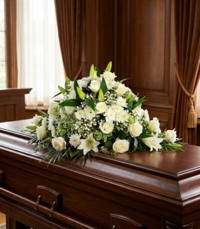 Cubre caja fúnebre solemne con rosas blancas, lirios y follaje verde sobre un ataúd de madera oscura en un entorno elegante y