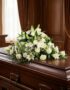 Cubre caja fúnebre solemne con rosas blancas, lirios y follaje verde sobre un ataúd de madera oscura en un entorno elegante y