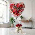 Globo metalizado en forma de corazón con la frase 'Te Amo' flotando sobre un arreglo de rosas rojas frescas en un jarrón de c