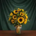 Jarrón de cristal transparente con un arreglo de 12 girasoles vibrantes, hojas verdes y pequeñas flores amarillas, atado con