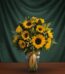Jarrón de cristal transparente con un arreglo de 12 girasoles vibrantes, hojas verdes y pequeñas flores amarillas, atado con