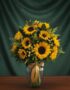 Jarrón de cristal transparente con un arreglo de 12 girasoles vibrantes, hojas verdes y pequeñas flores amarillas, atado con